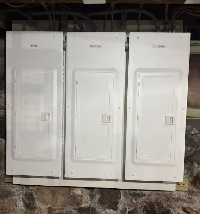 Three white electrical panels mounted on a stone wall in an unfinished utility room