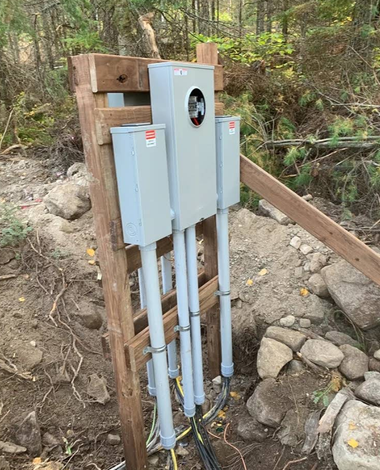 Three gray electrical meter boxes mounted on a wooden post beside a wooded dirt path.