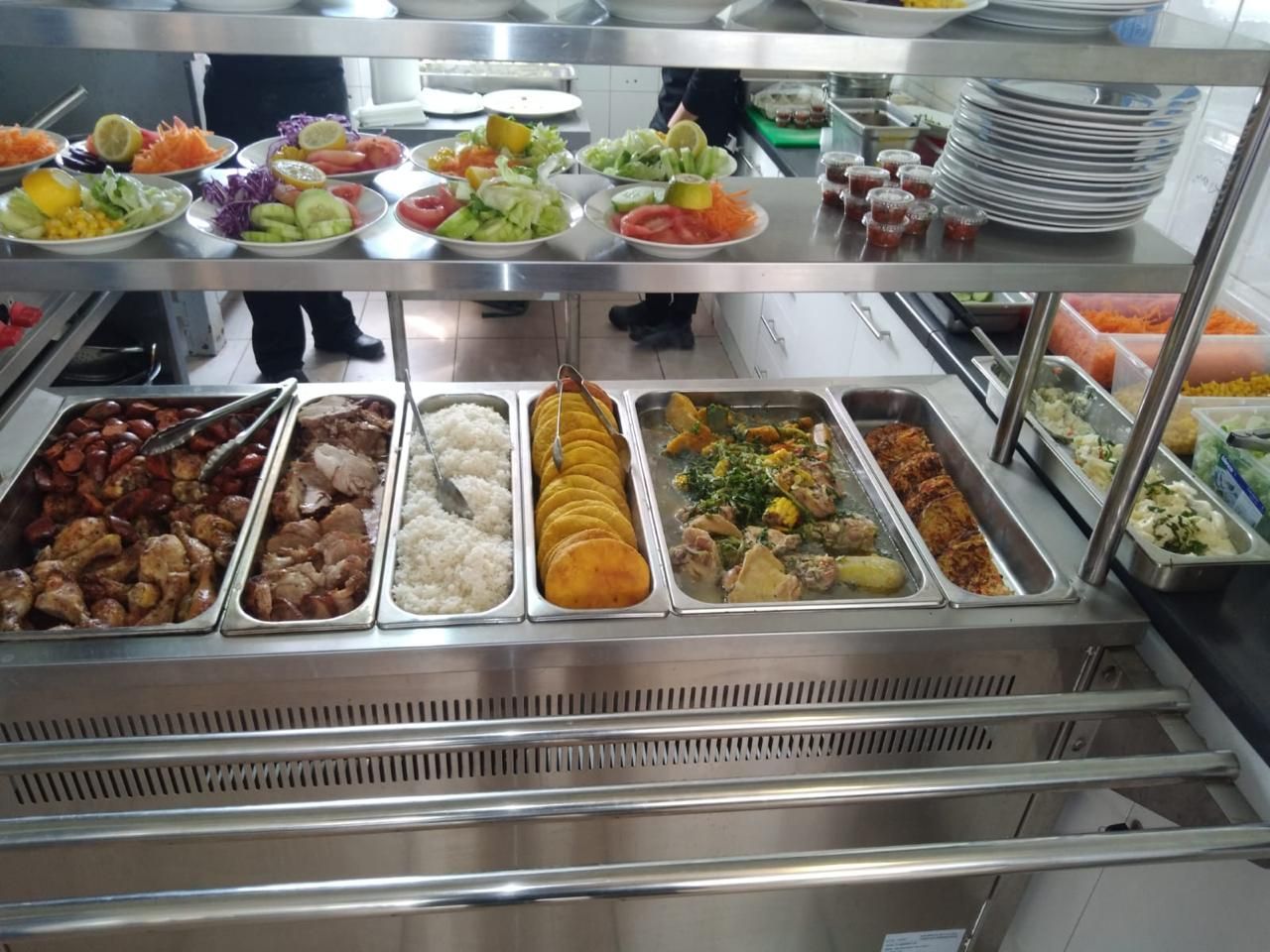 Buffet con diversos platos: carnes, arroz, verduras, ensaladas y guarniciones. Mostradores de acero inoxidable, 