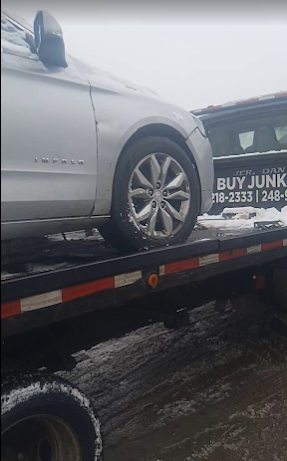 Silver car loaded on a tow truck in a rainy lot, with a partially visible “BUY JUNK” sign.
