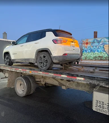 White SUV strapped on a flatbed trailer on a wet road under a blue sky.
