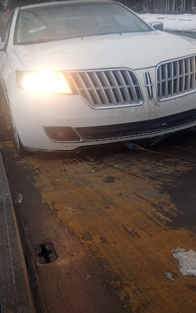 White luxury sedan with headlights on, parked on a wet tow truck bed at night