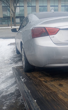 Silver sedan parked on a flatbed tow truck in a snowy lot near a building.