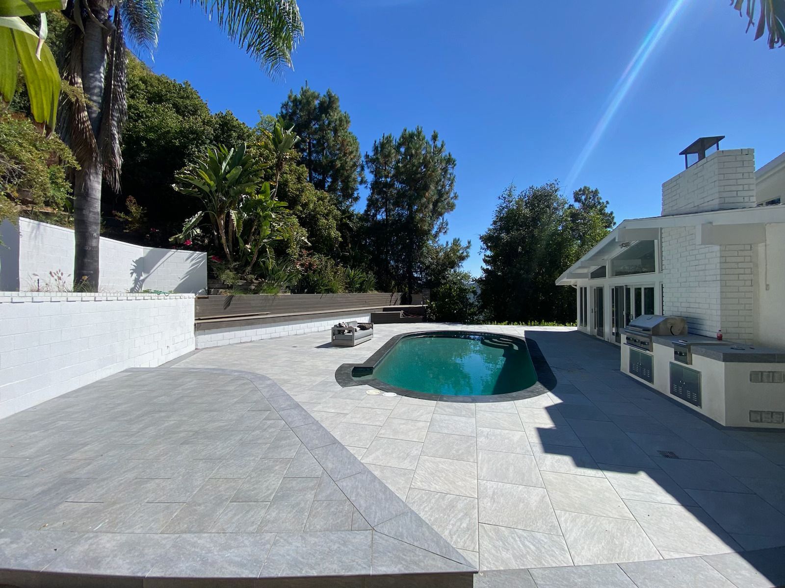 Sunny backyard patio with a turquoise pool, white tiled deck, and lush trees around a modern house