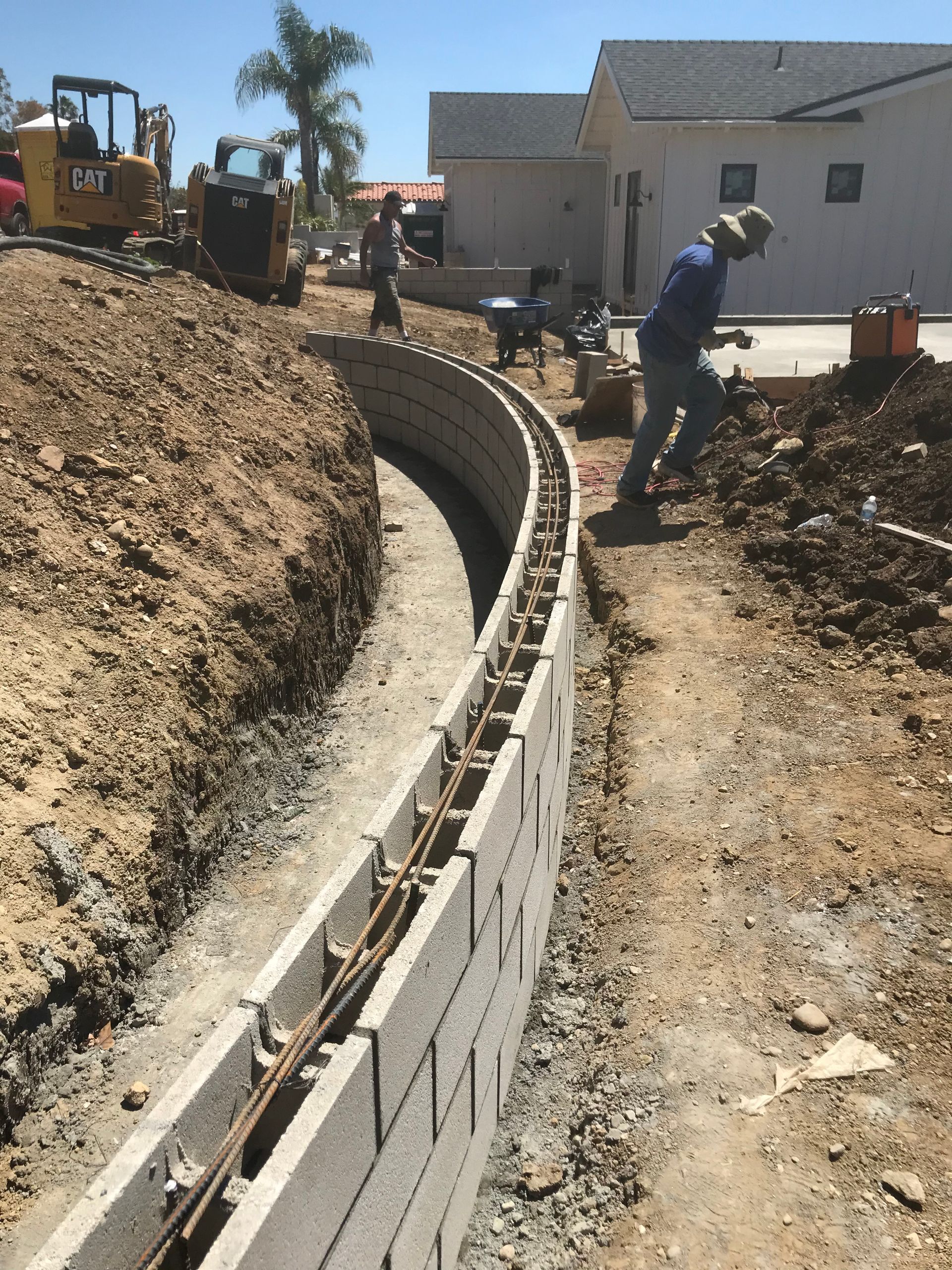 Workers building a curved cinder block retaining wall beside a dirt trench and house under construction