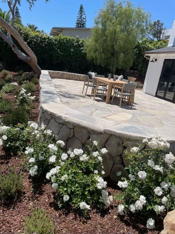 Stone patio with wooden table and chairs, surrounded by white flowers and greenery in a sunny garden