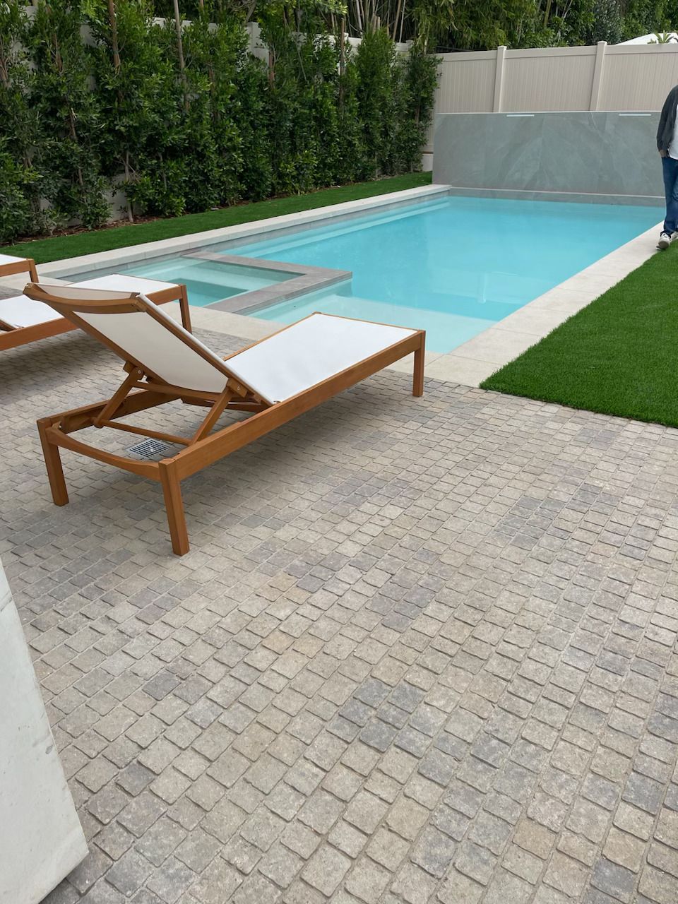 Modern backyard pool with two wooden lounge chairs on a stone patio, bordered by hedges and green lawn
