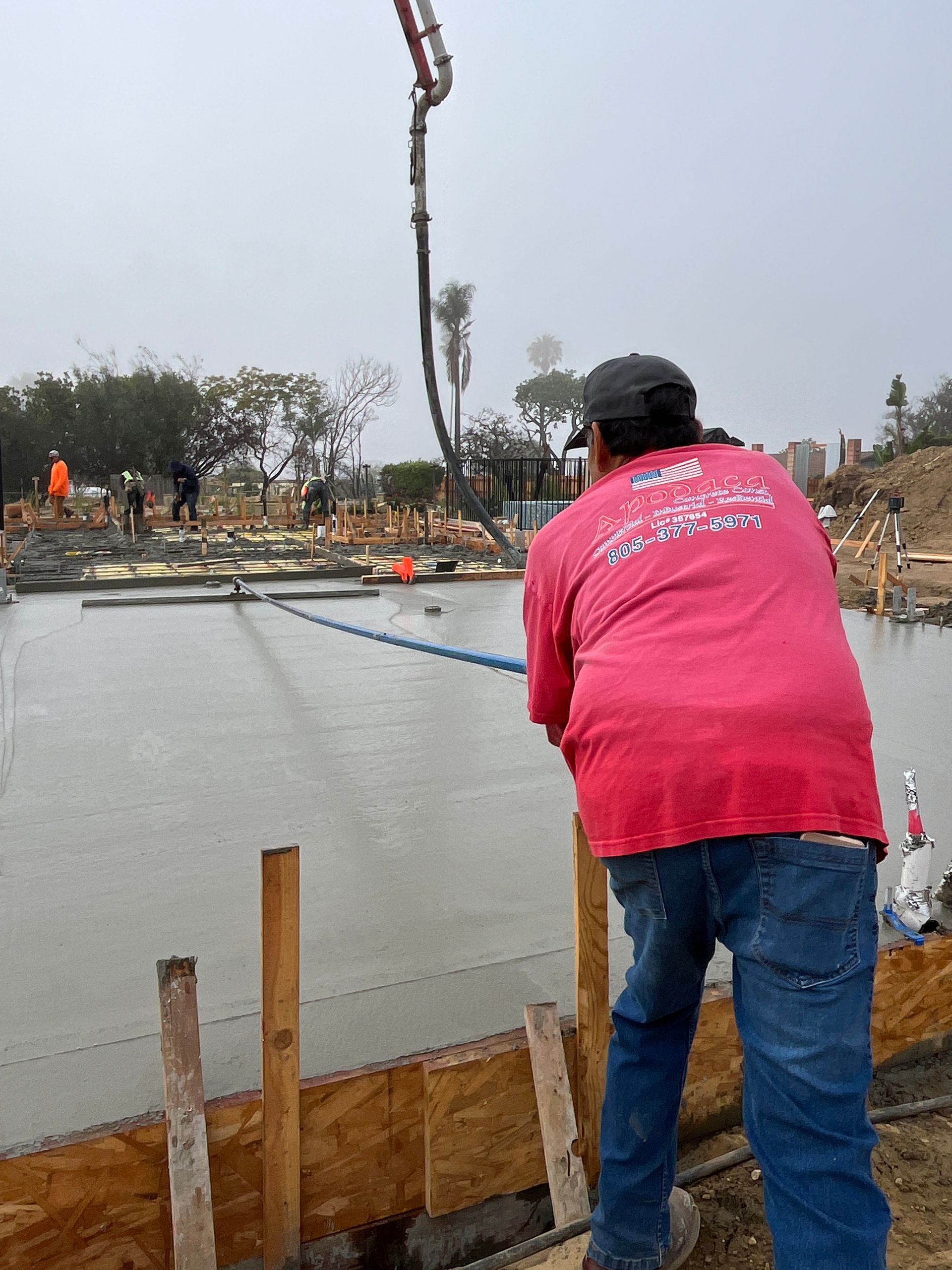Worker in a red shirt pouring concrete beside a foundation at a construction site.