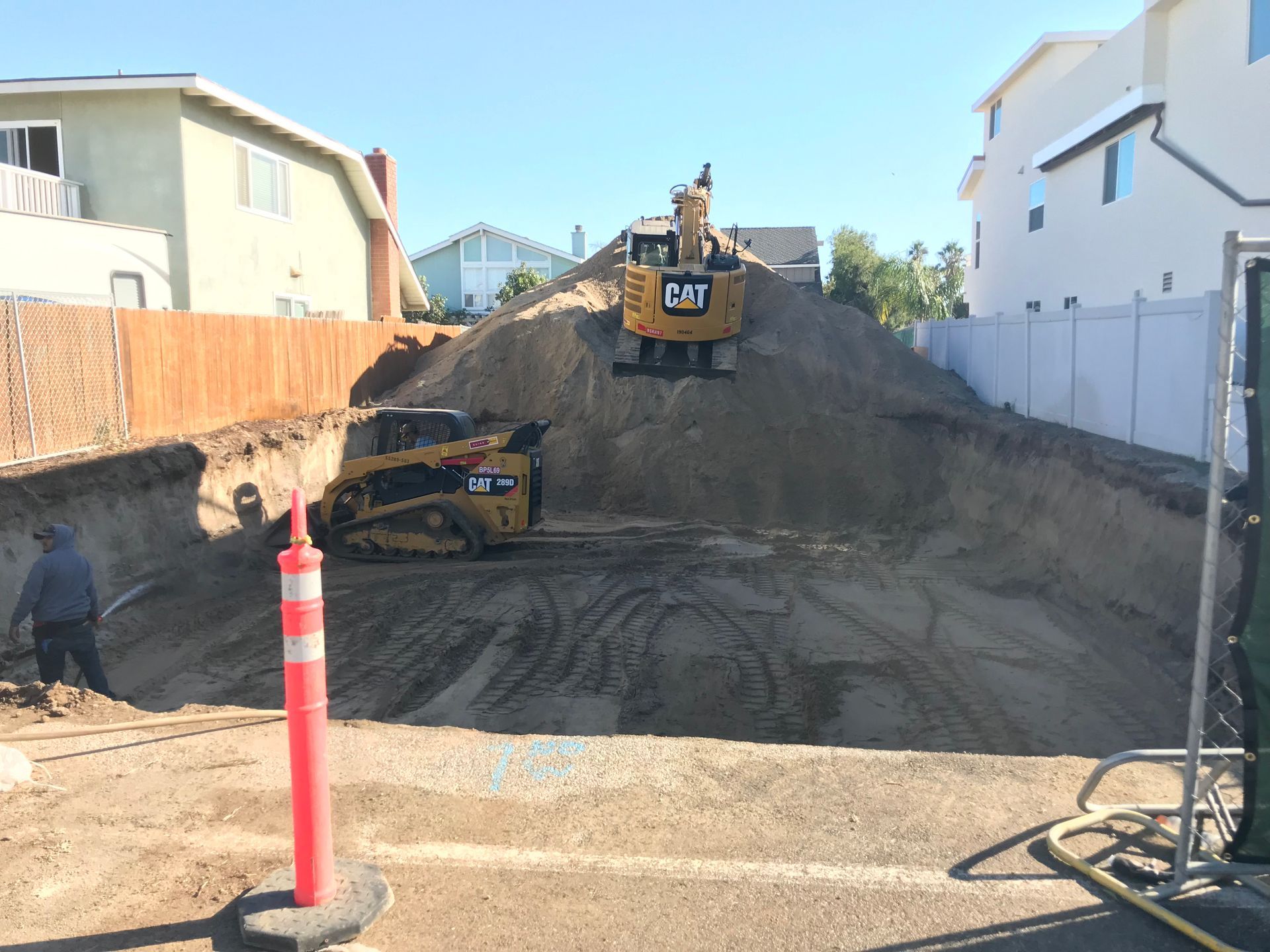 Construction site with excavator on a dirt lot between houses, red post in foreground.