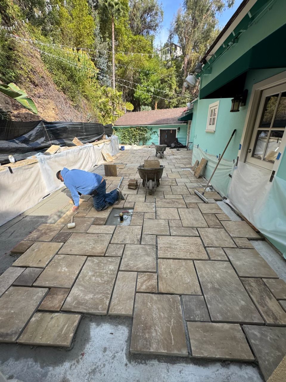 Worker laying stone pavers on a backyard patio beside a turquoise house