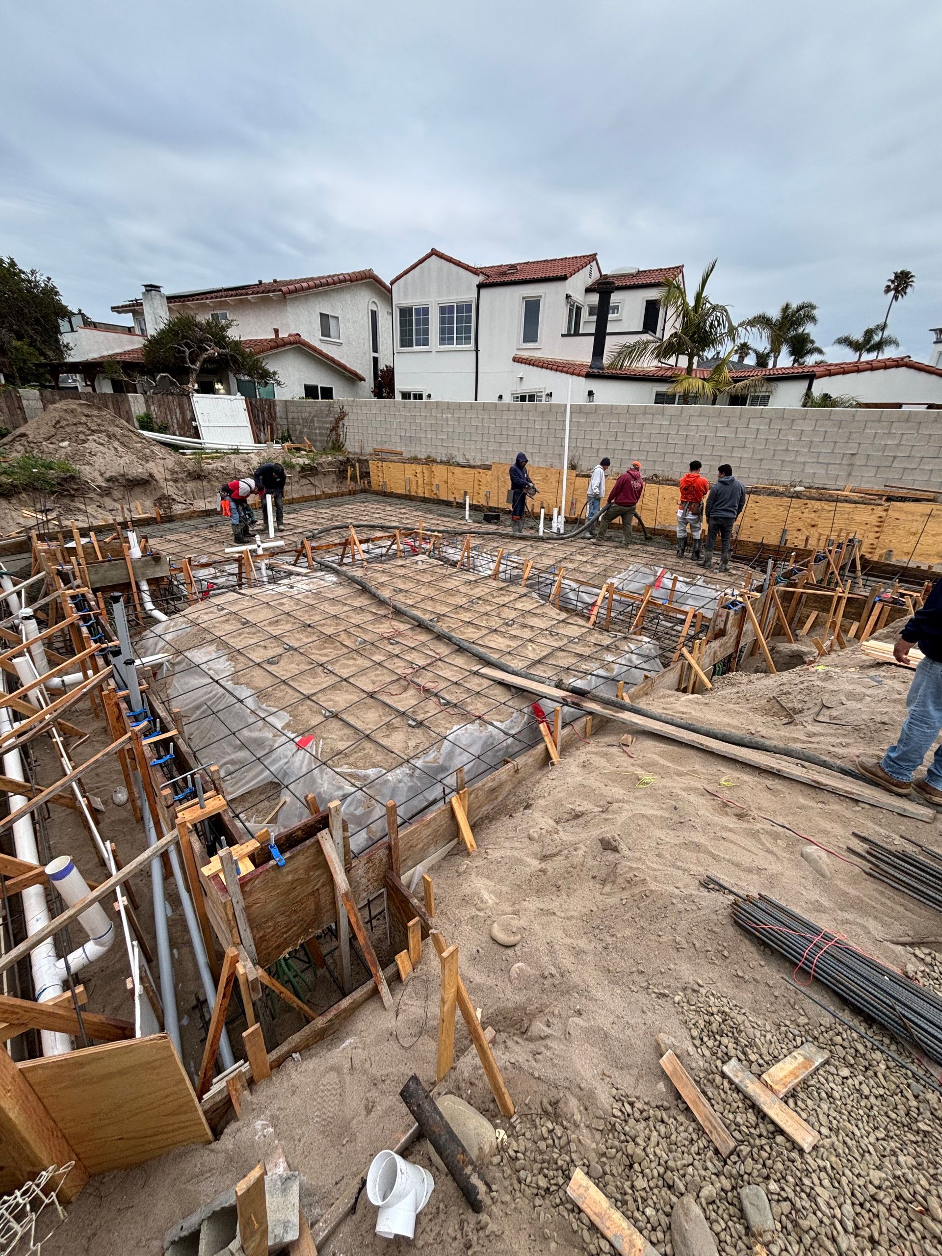 Construction site with workers reinforcing a foundation, rebar grid, and white houses in the background