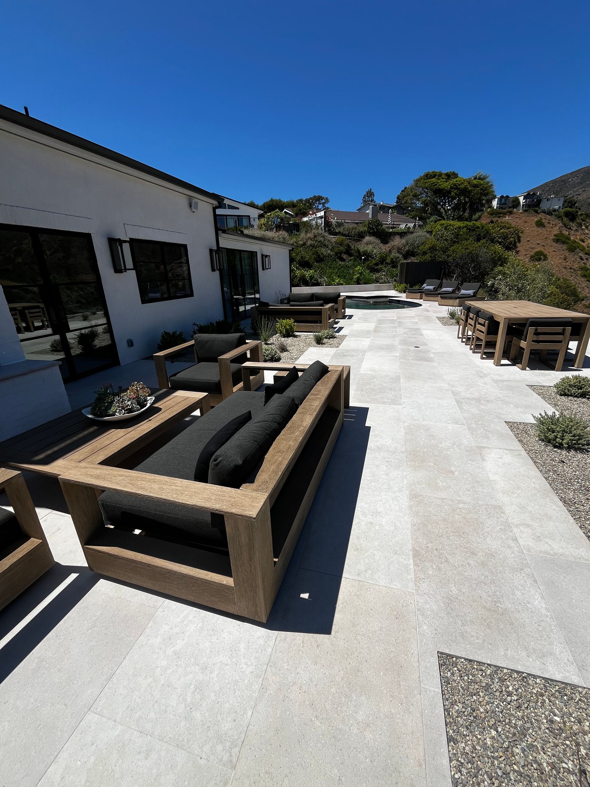 Sunny backyard patio with gray furniture, concrete deck, and hillside view beside a white house