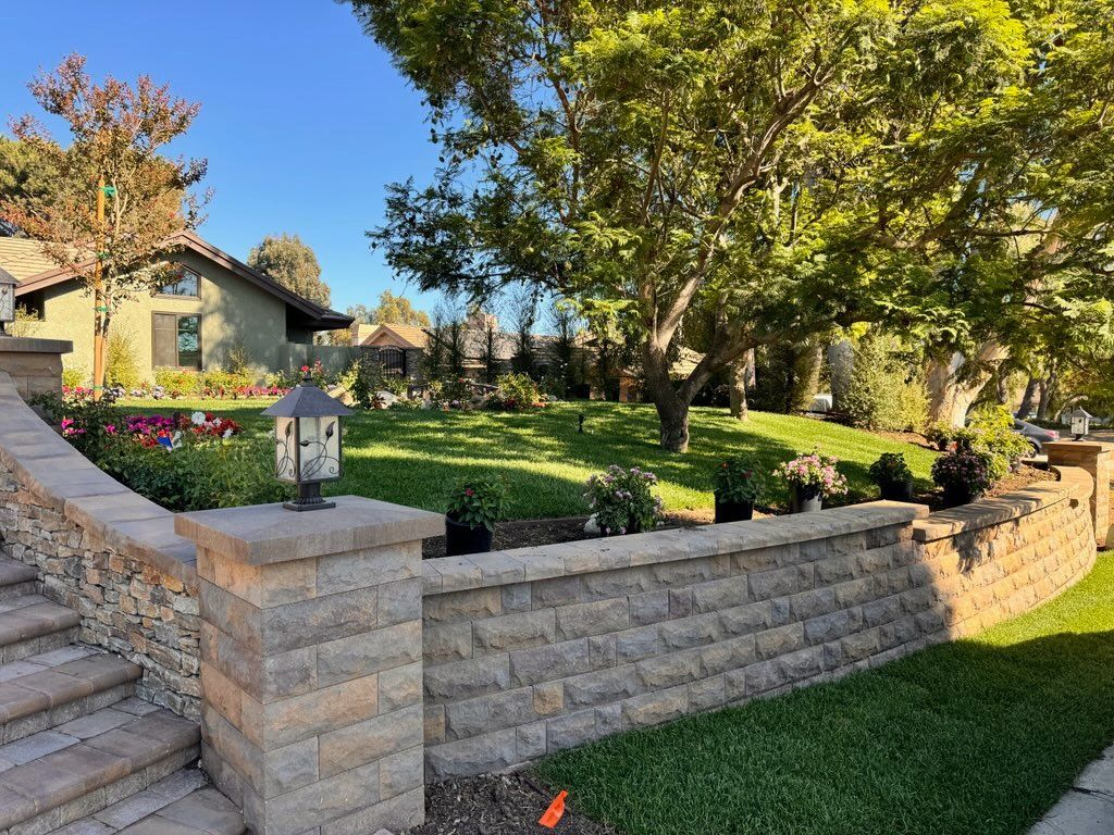Landscaped front yard with stone retaining walls, stairs, green lawn, trees, and a house in the background