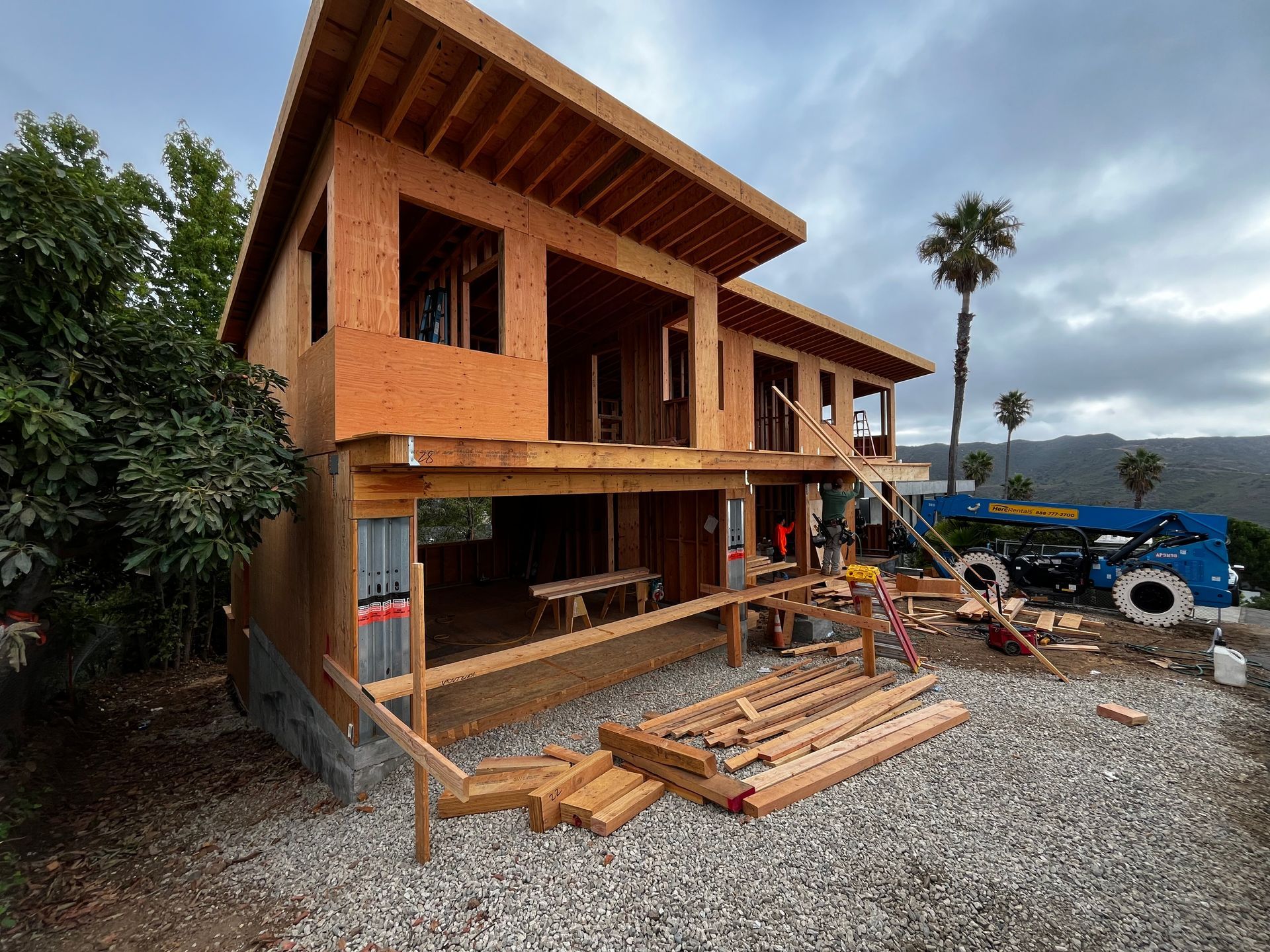 Two-story house under construction with exposed wood framing, gravel yard, and construction equipment nearby
