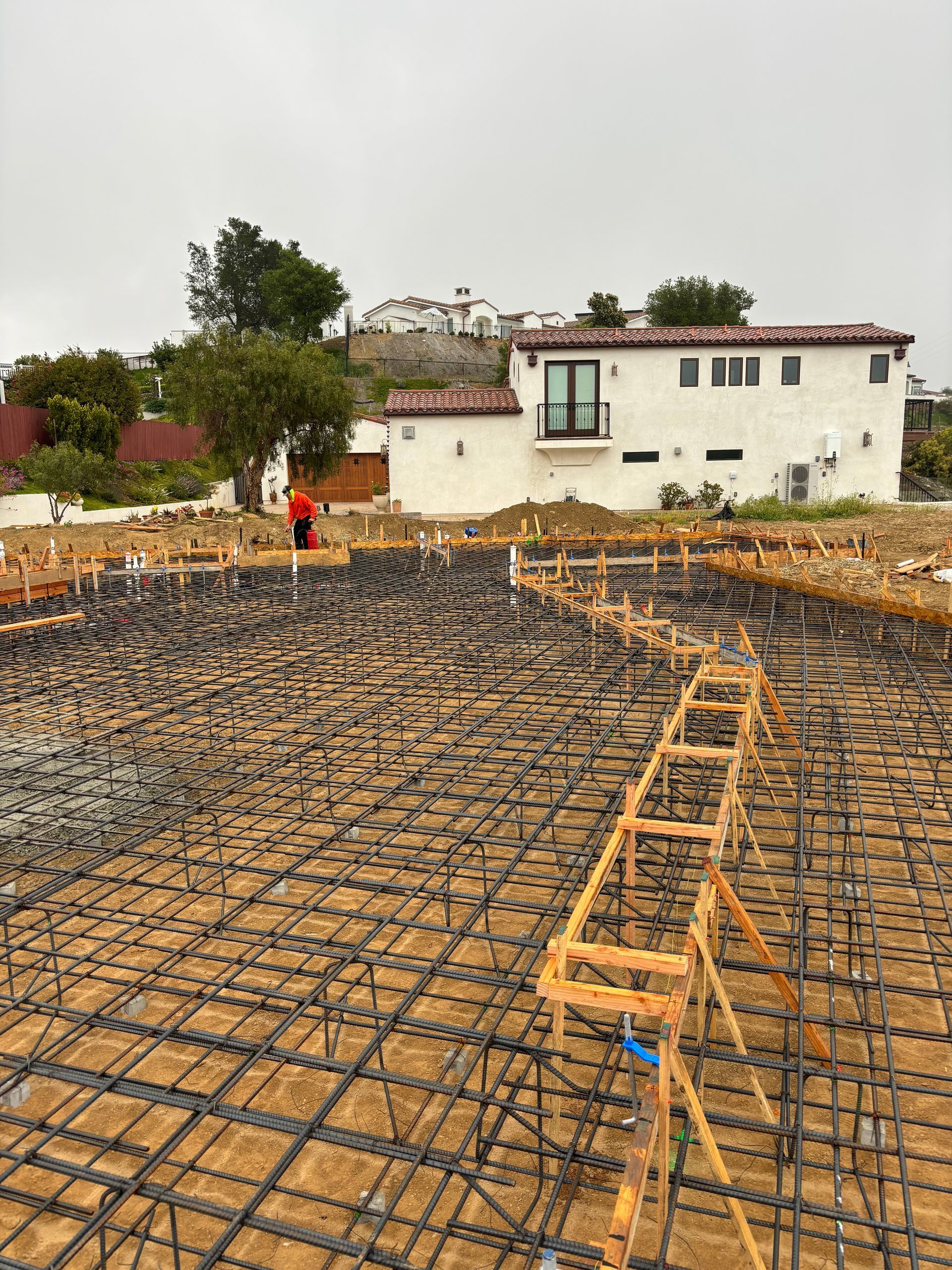 Construction site with rebar foundation grid and concrete forms in front of white houses and trees