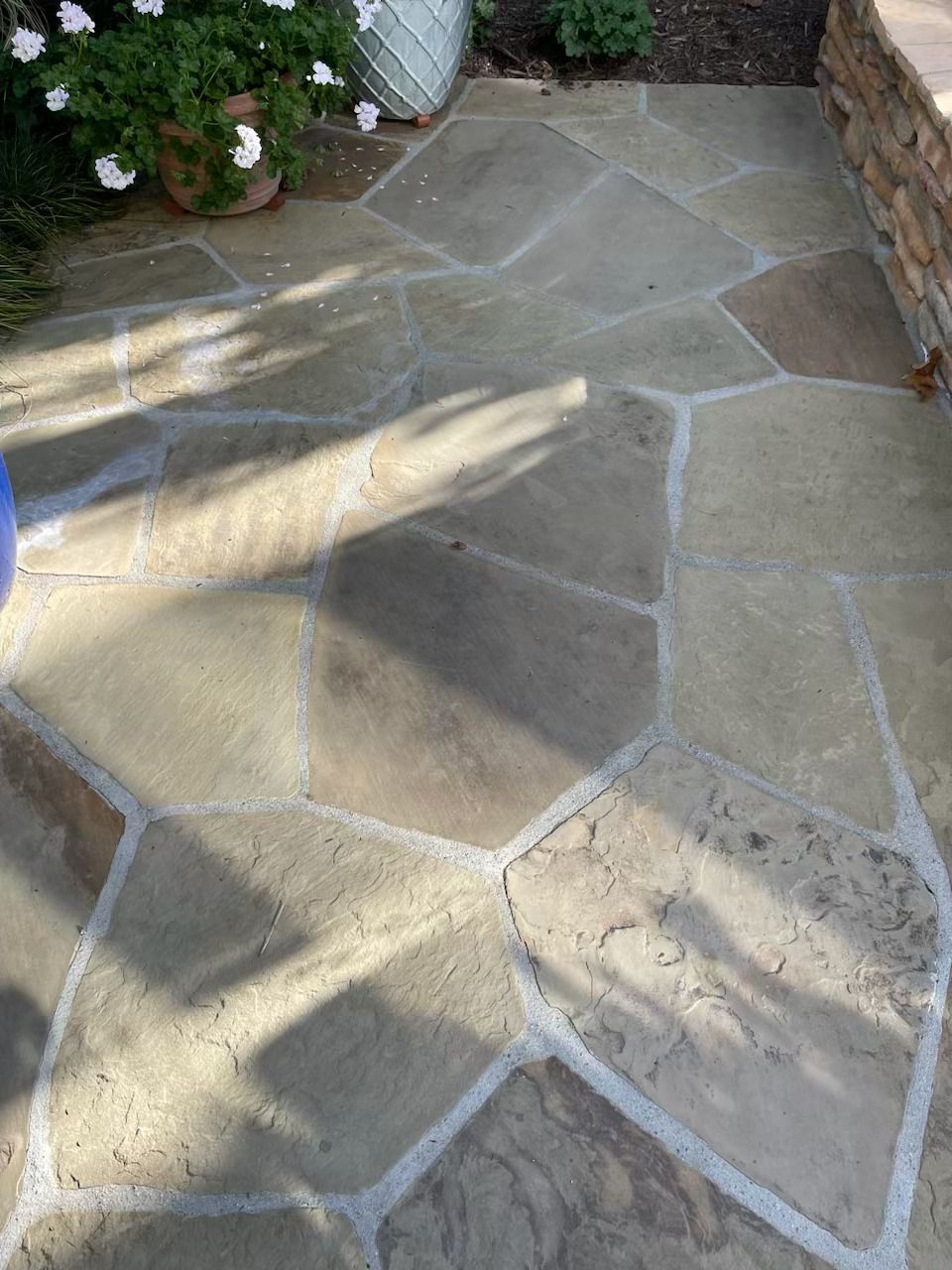 Stone garden walkway with irregular tan paving stones and sunlight shadows near plants and a brick wall