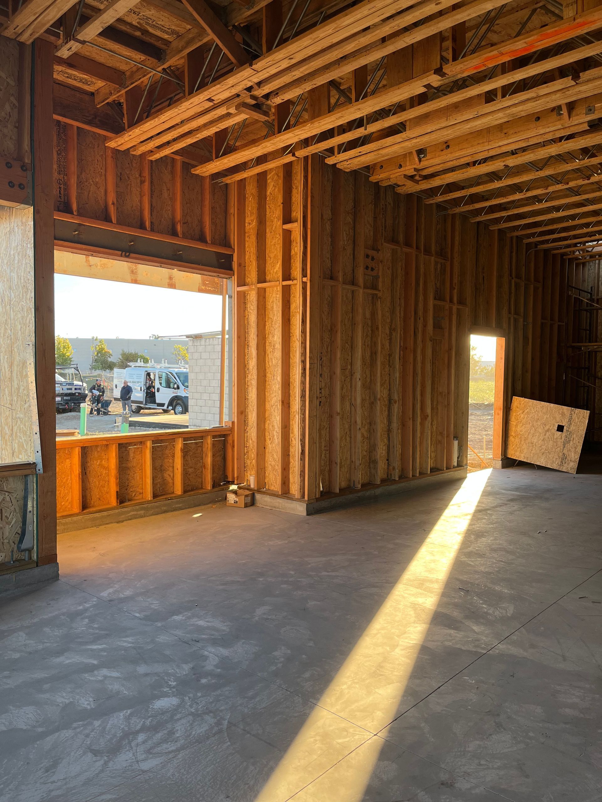 Unfinished wooden interior framing with sunlight streaming through window openings onto a concrete floor.