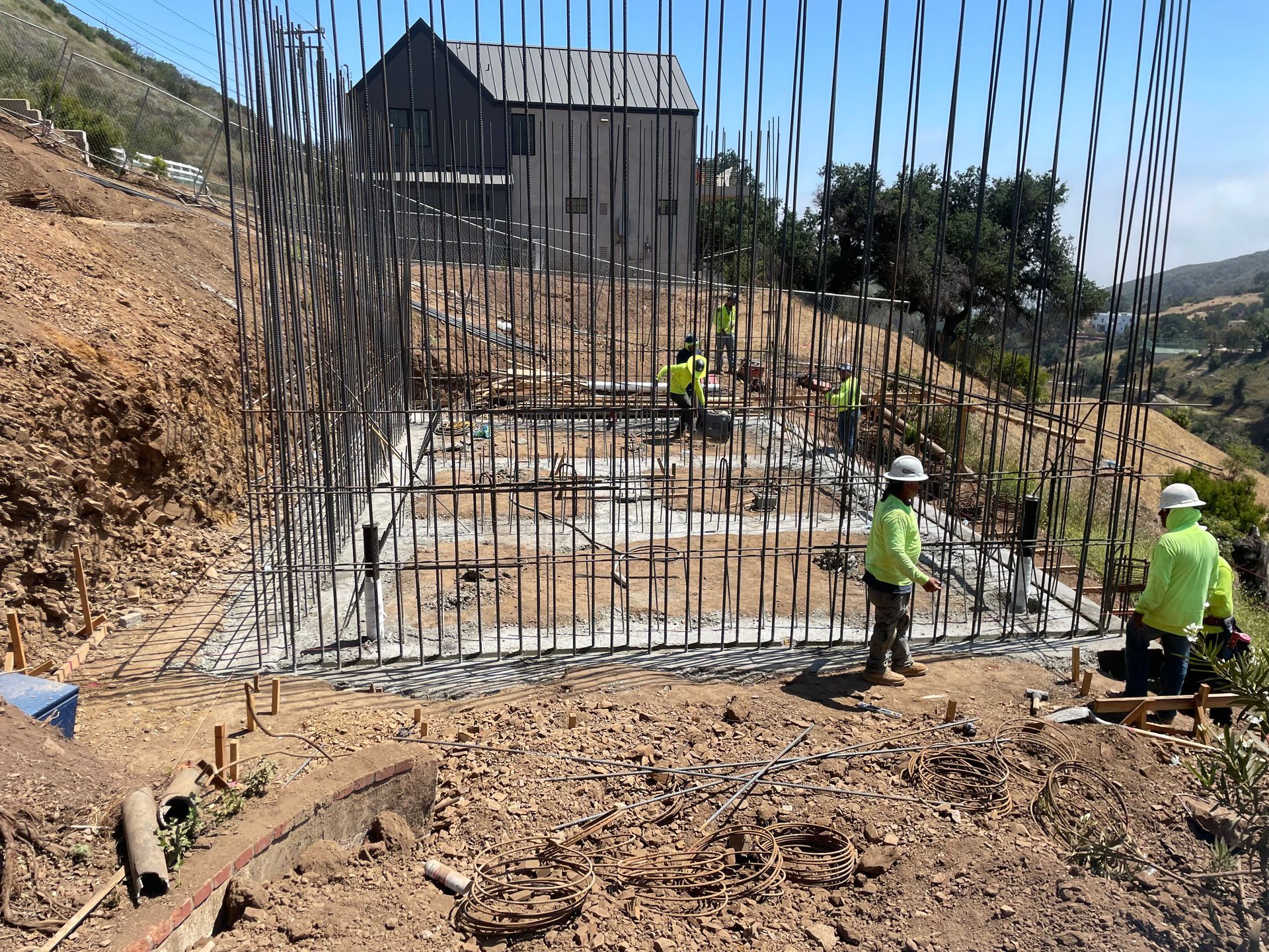 Workers pouring concrete foundation on a hillside construction site beside exposed rebar columns
