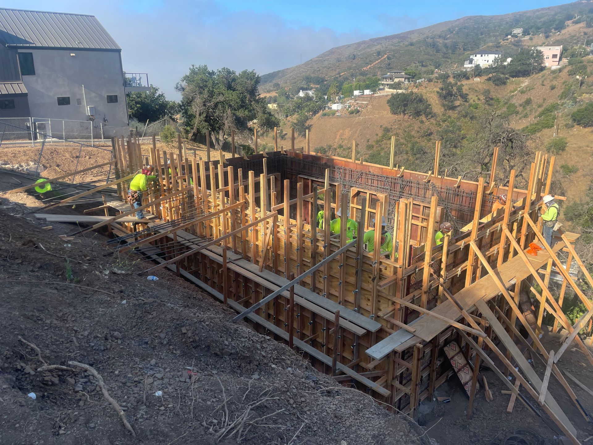 Construction site with wooden foundation forms and workers on a hillside near houses