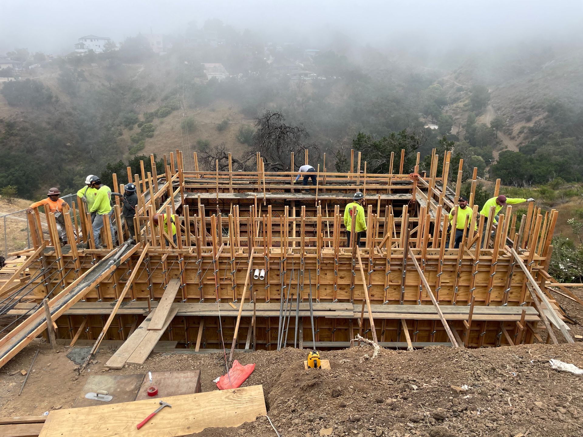 Construction workers assemble wooden formwork on a concrete foundation in a foggy outdoor site.