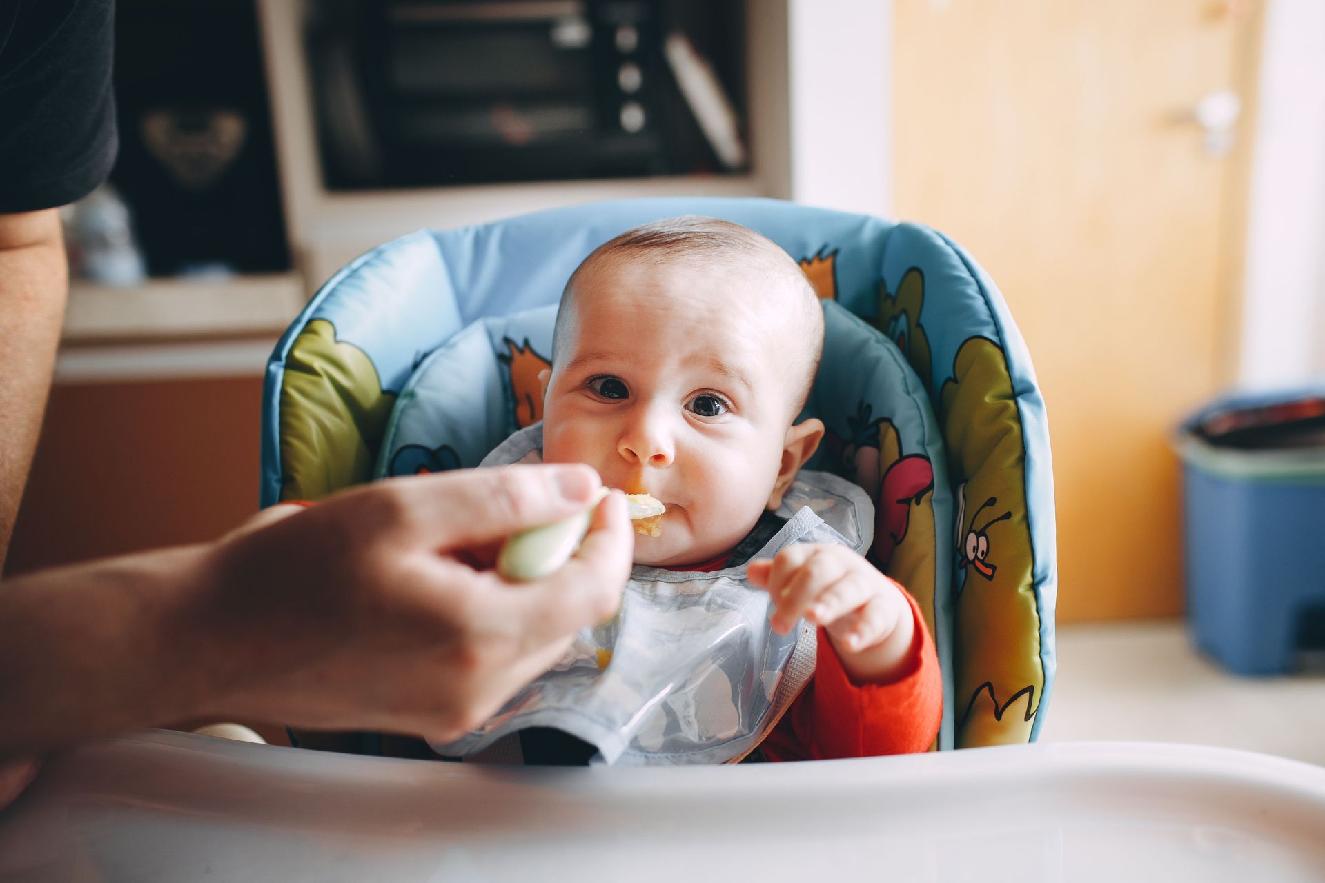 Baby in a high chair being fed with a spoon, looking at the camera.