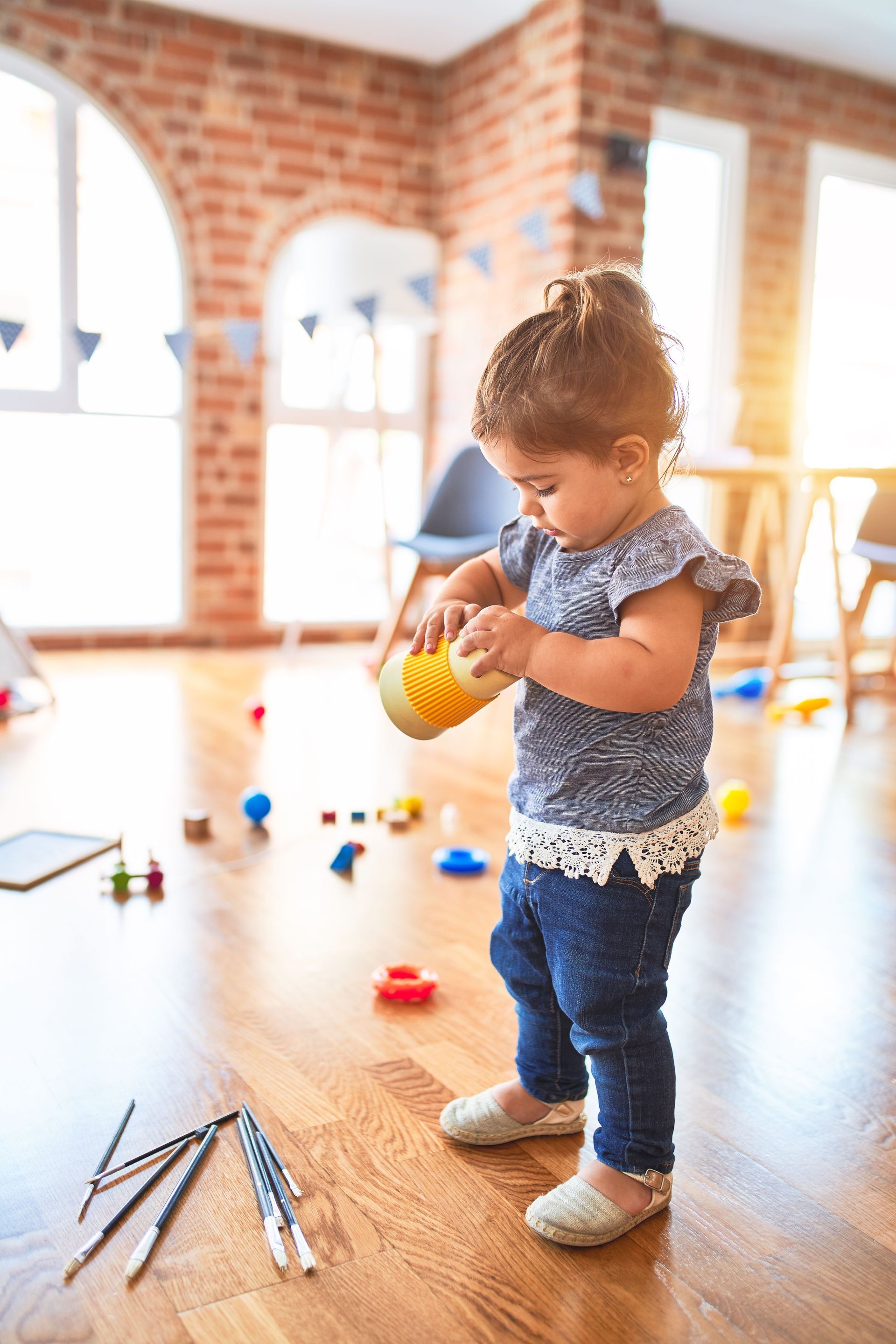 Toddler stacking yellow cups on a wooden floor in a bright playroom with scattered toys.