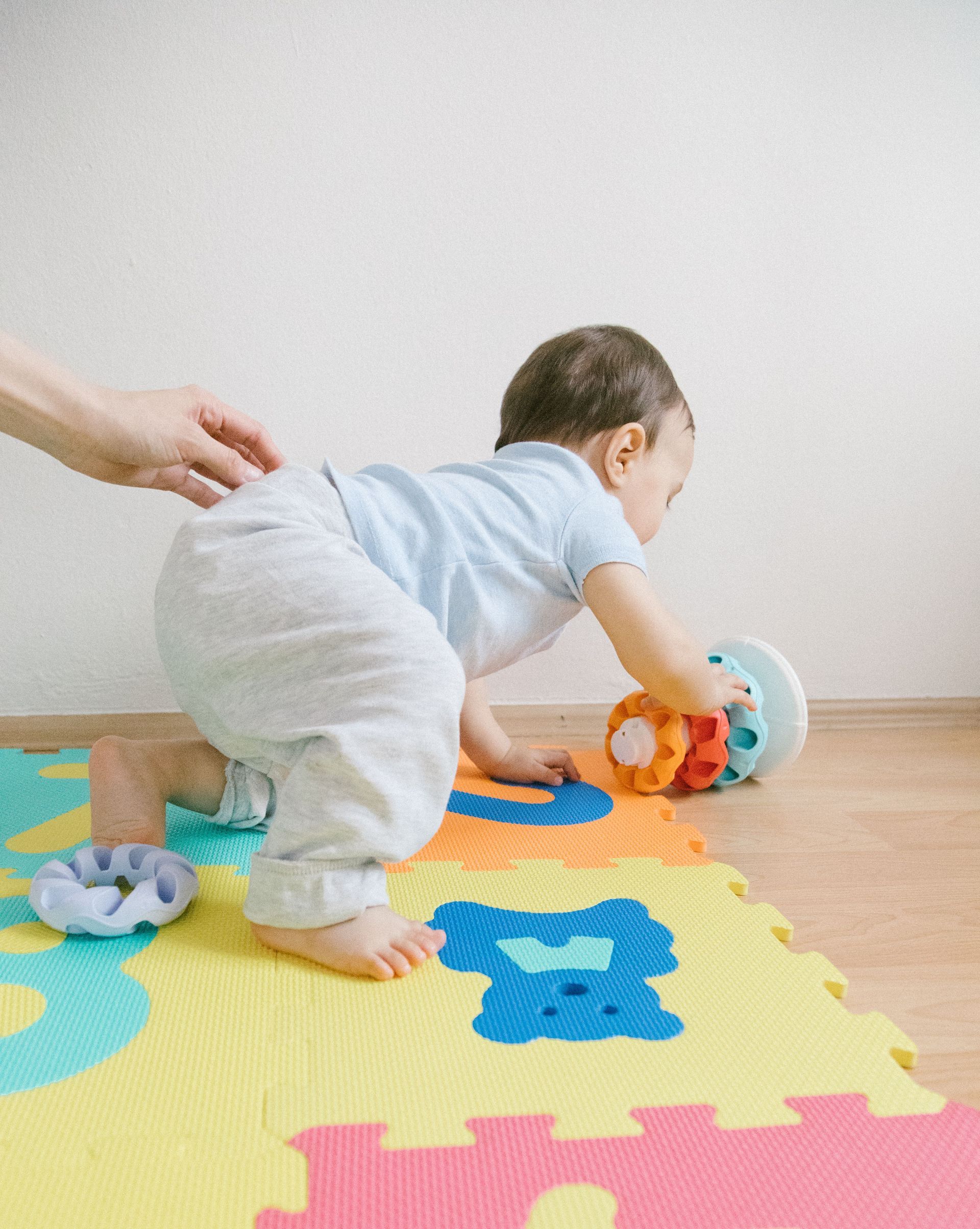 Baby crawling on colorful foam mat with a hand nearby and toys on a wooden floor.