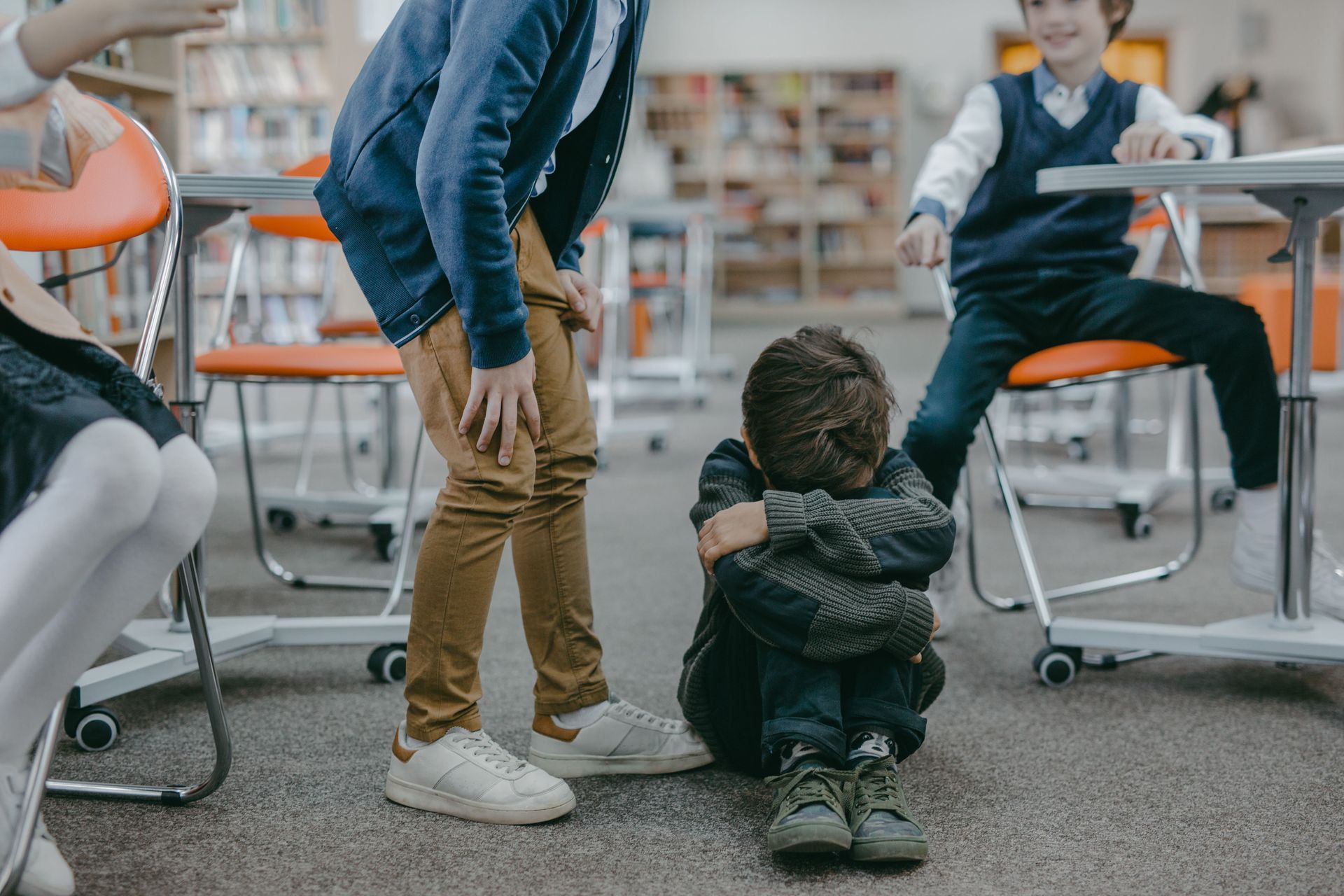 Child sitting on classroom floor while an adult bends down nearby and other students sit at desks