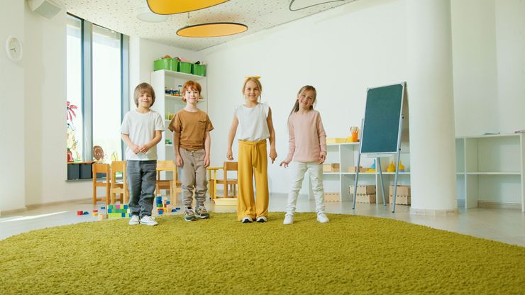 Four children standing in a bright classroom with a green rug, shelves, and a chalkboard.