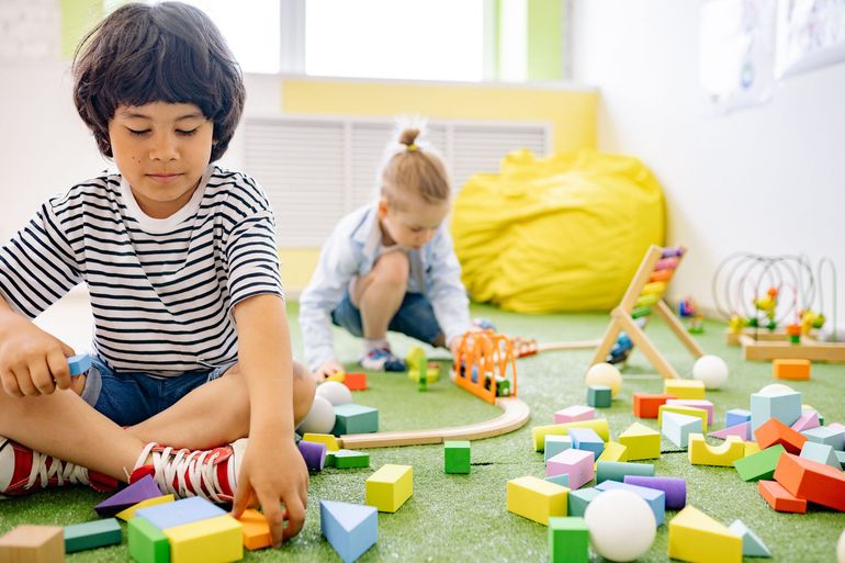 Children playing with colorful building blocks on a green carpet in a bright playroom.