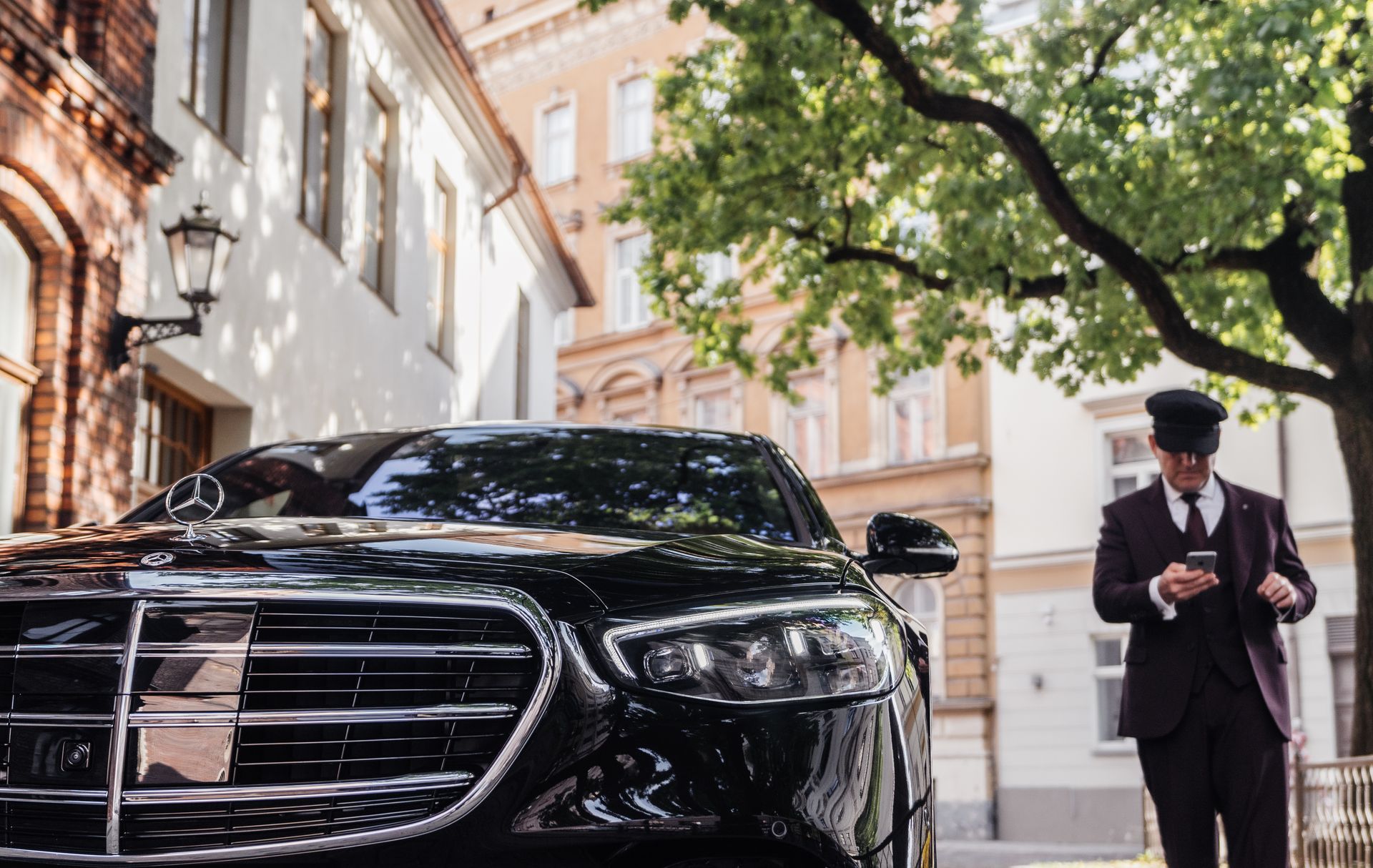 Black luxury car parked on a city street beside a doorman in a suit checking his phone
