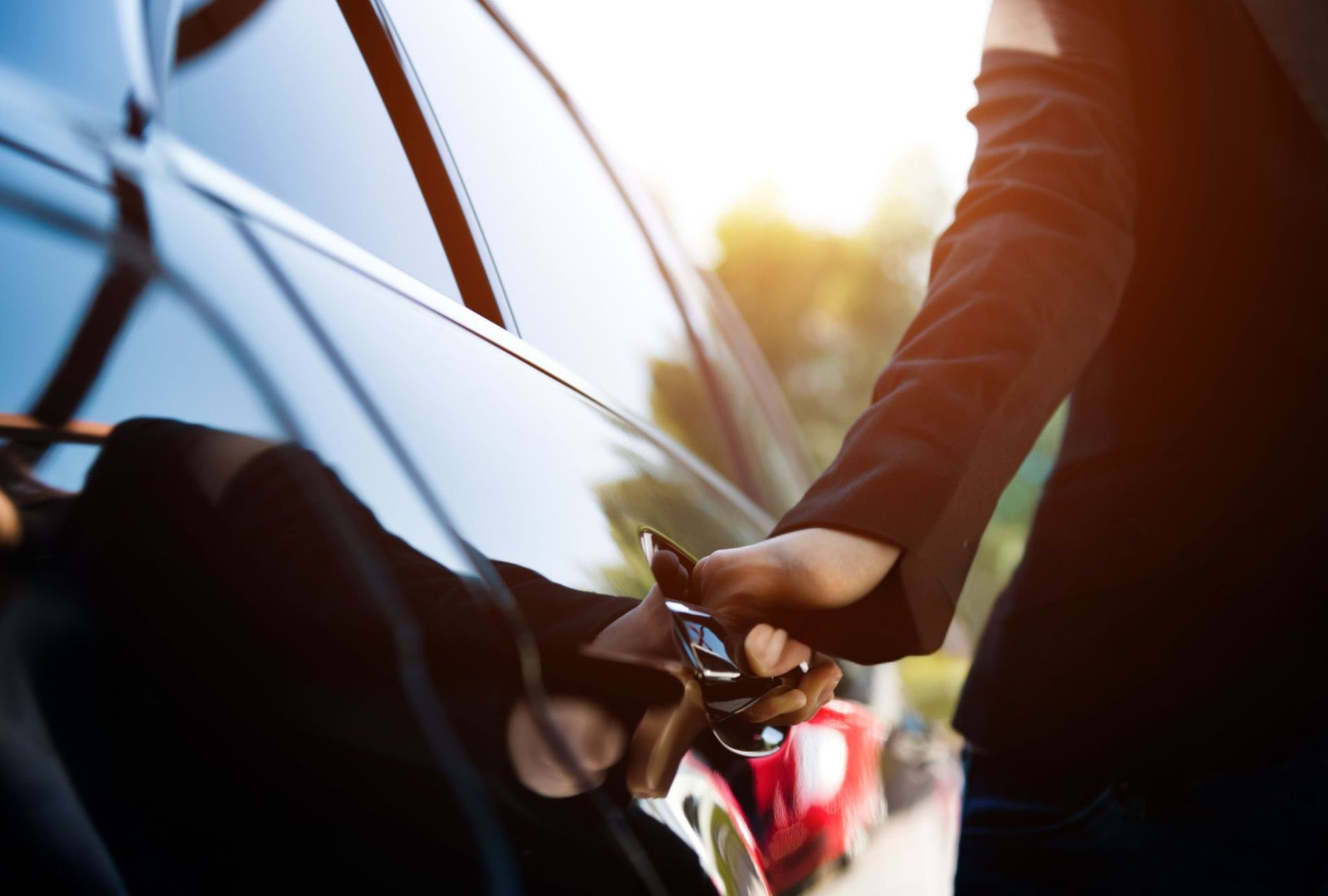 Person opening a black car door in warm sunlight, with other cars blurred nearby