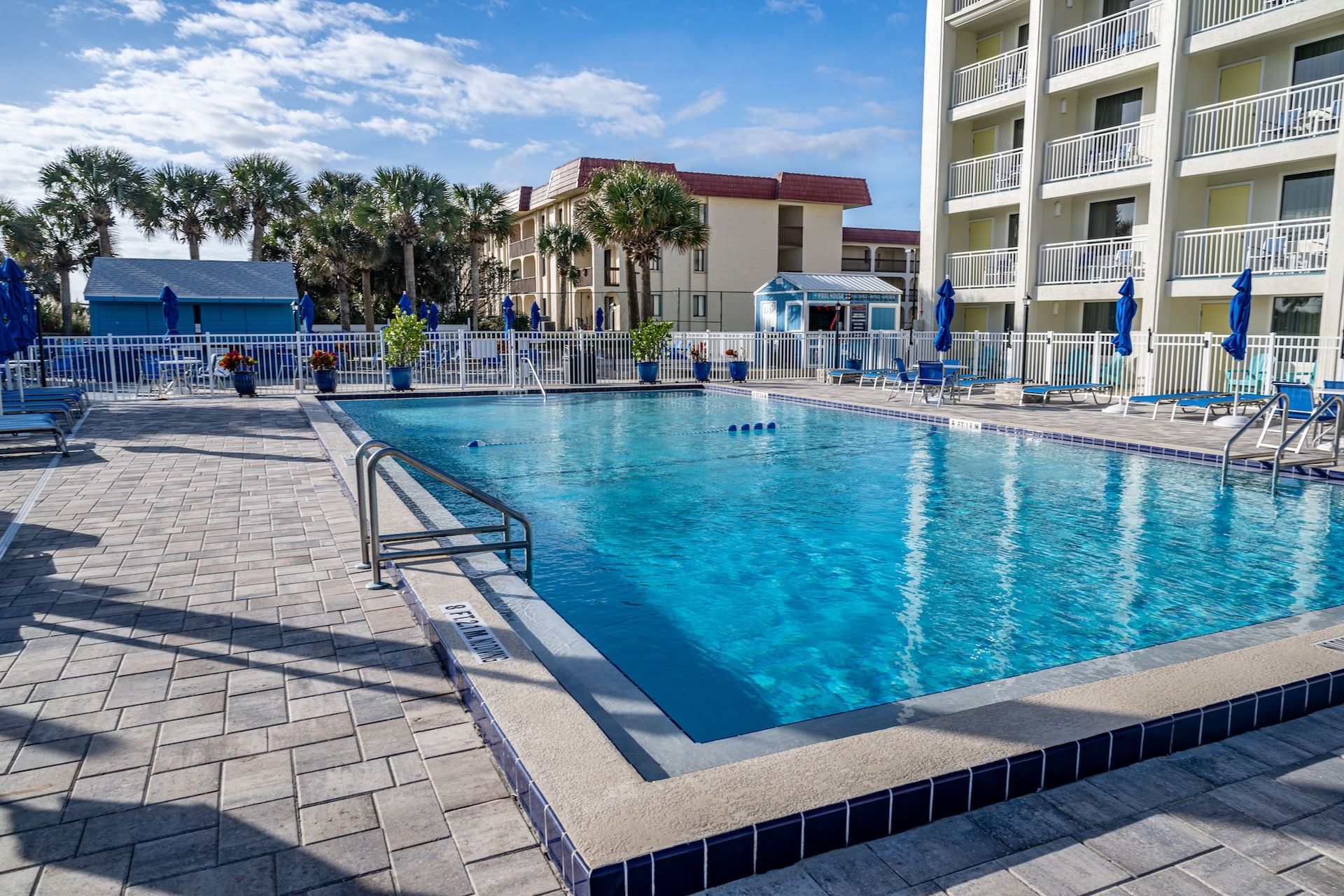 Outdoor resort pool with blue water, lounge chairs, umbrellas, and palm trees under a sunny sky