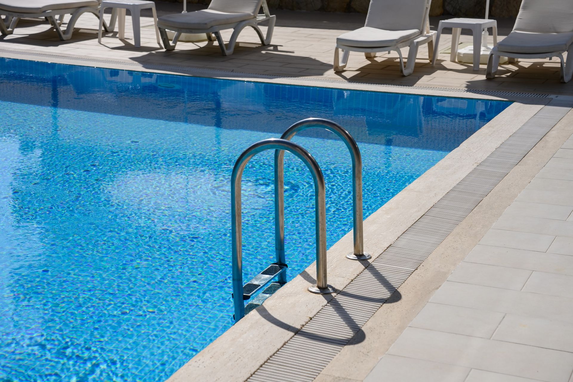 Swimming pool ladder at the edge of a blue tiled pool with lounge chairs in the background