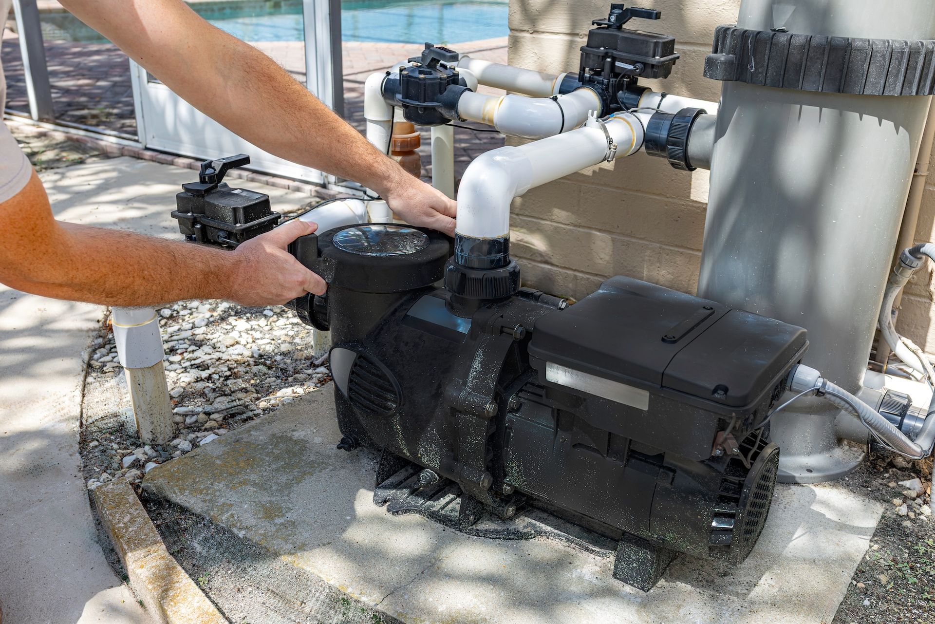 Person adjusting pipes on a black pool pump beside outdoor equipment and gravel ground