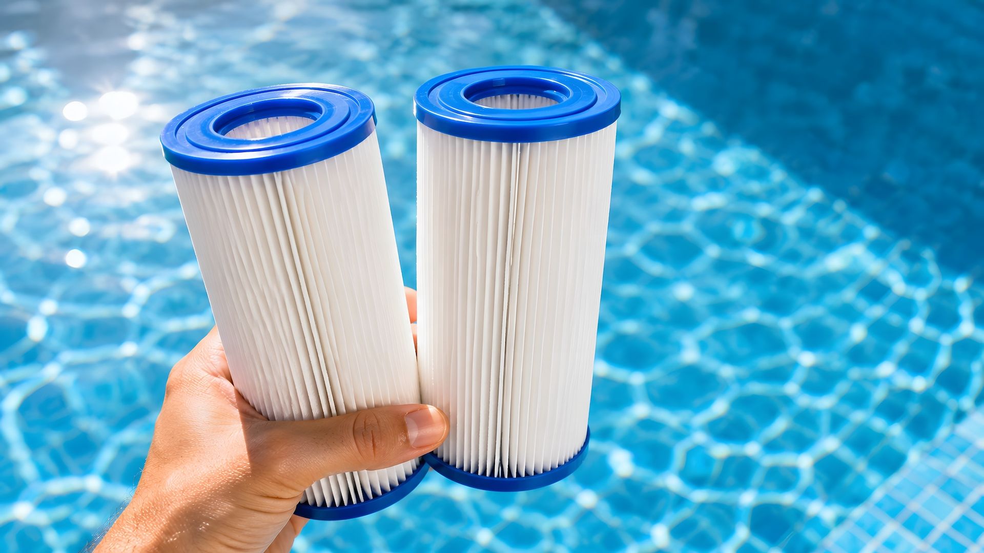 Hand holding two blue-and-white pool filter cartridges over a swimming pool background