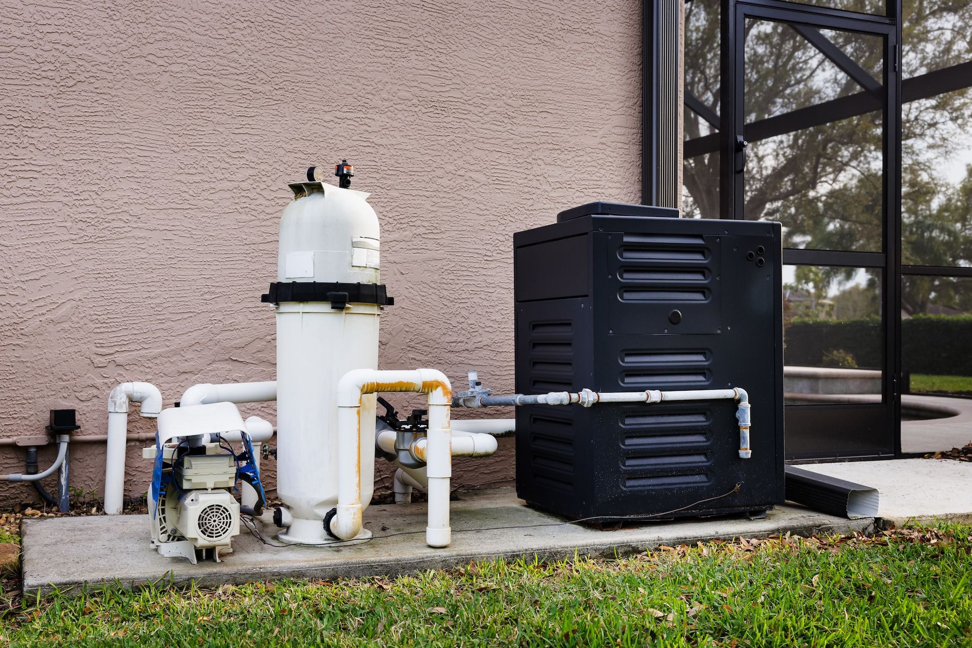 Outdoor pool equipment with pump, white filter tank, and black heater beside a house wall