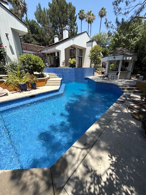 Backyard swimming pool with blue tile, white patio, and trees in bright sunlight
