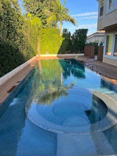 Backyard swimming pool with a built-in circular spa beside a house, bordered by hedges and palm trees.