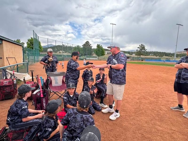Youth baseball team in red and black uniforms huddling, hands in the center, smiling and celebrating outdoors.