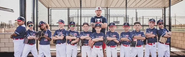 Youth baseball team posing with arms crossed; they wear white pants, navy tops and caps, against a chain link fence.