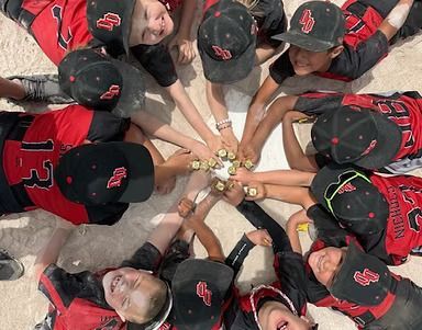 Youth baseball team in red and black jerseys and hats, huddled together on a field, smiling and touching hands in a circle.