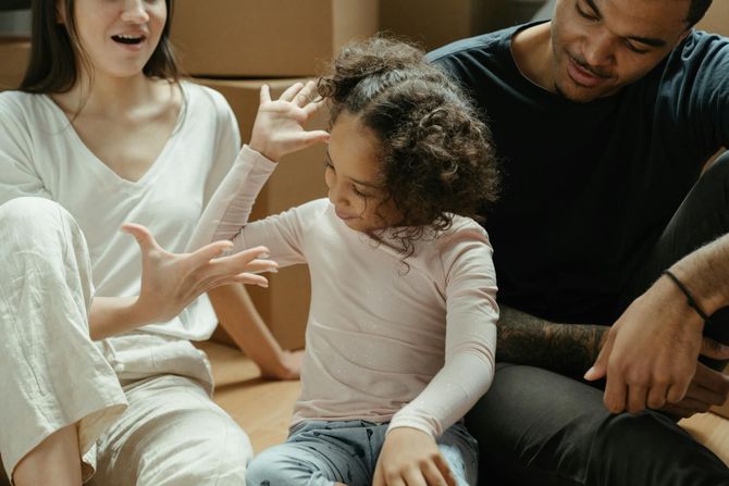 A young child sitting between two adults, playing a hand-slapping game, with cardboard boxes in the background.