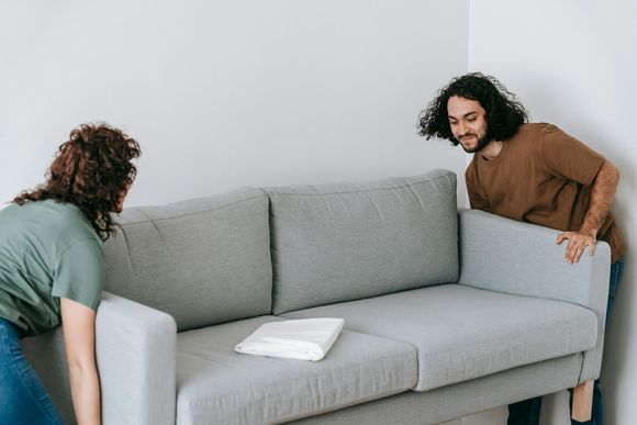 Two people move a light gray sofa against a plain white wall.