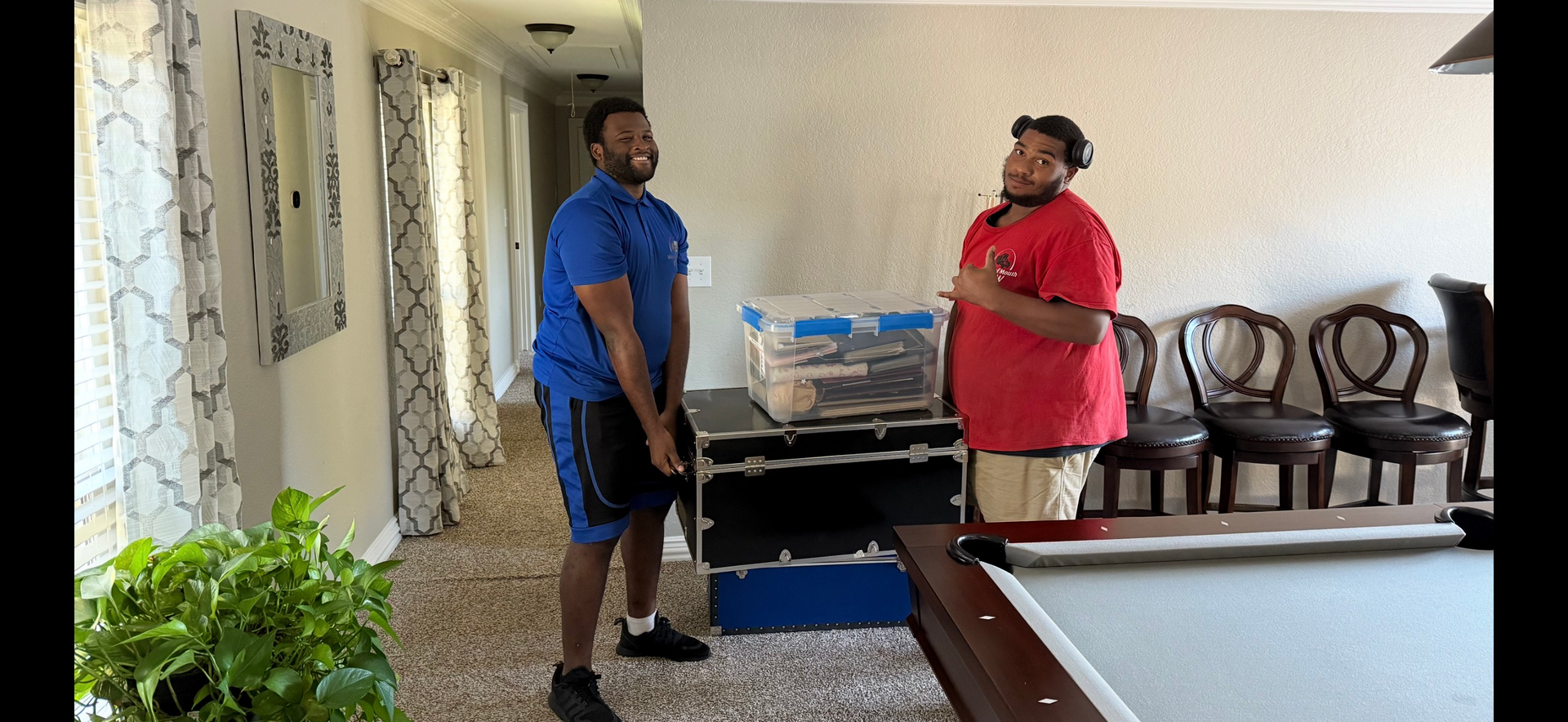 Two men in a hallway; one in blue shorts holds a black case, the other in red shirt gestures towards a pool table.