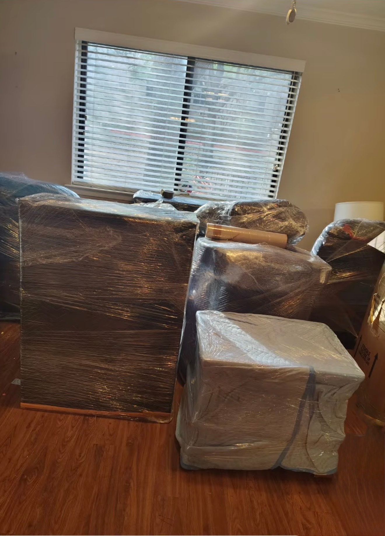 Boxes wrapped in plastic are stacked in a room with a window and closed blinds.