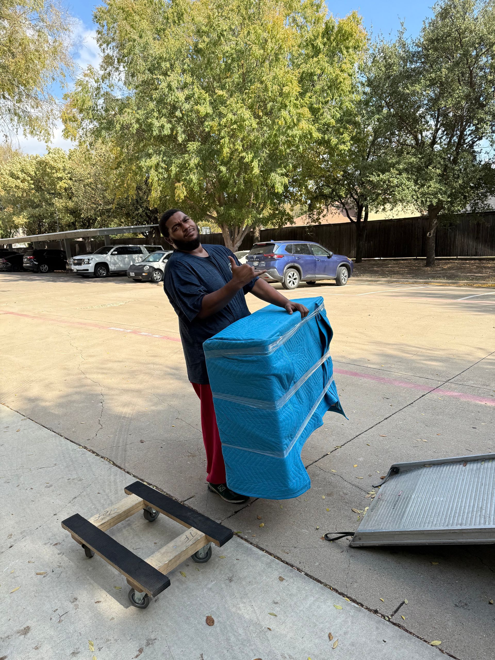 Person lifting a large, blue-wrapped item, next to a dolly. Outdoors, sunny.