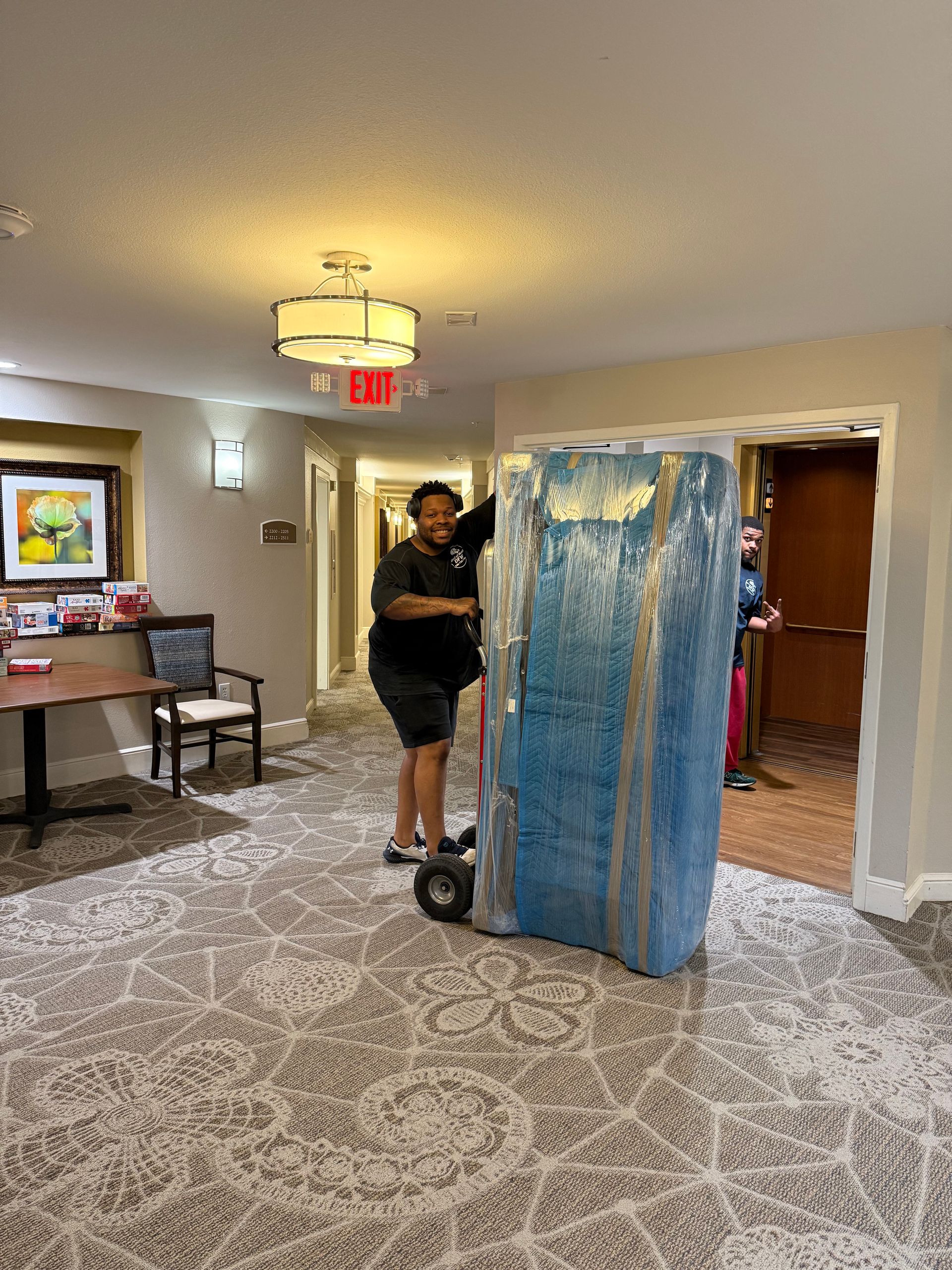 Person moving a large, blue-wrapped object on a dolly down a hallway with a patterned carpet.