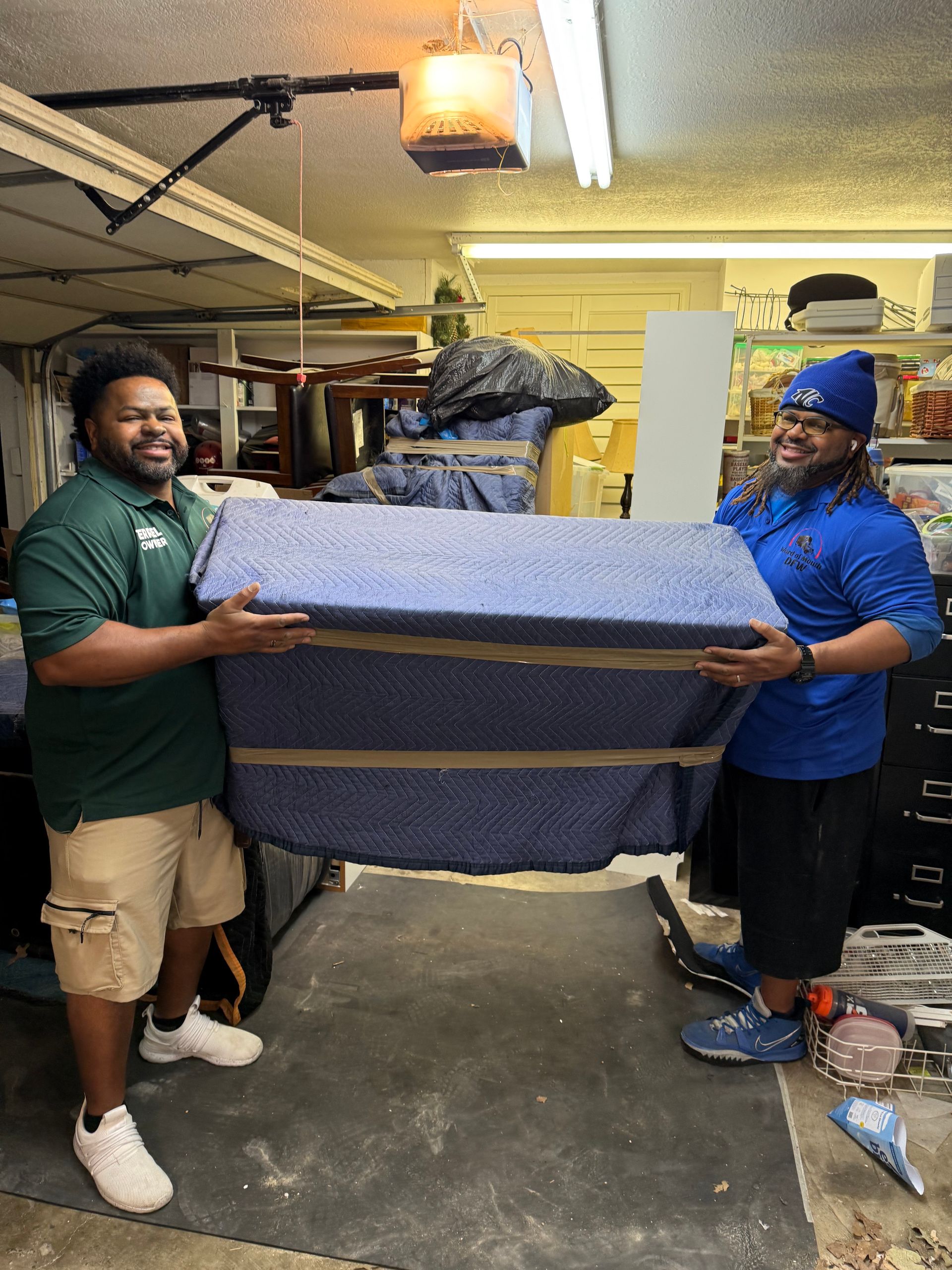 Two people carrying a large, blue furniture piece wrapped in padding inside a garage.
