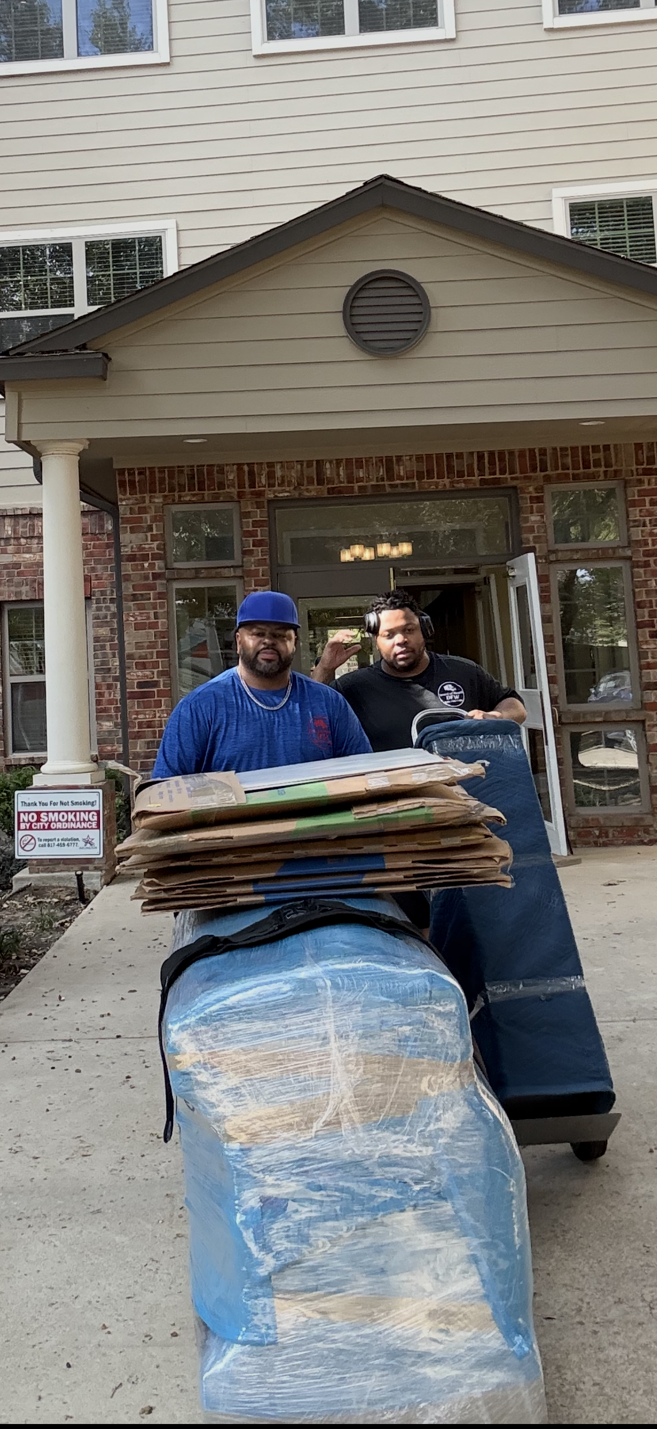 Two people moving furniture out of a brick building. One carries cardboard, the other pushes a dolly with wrapped items.